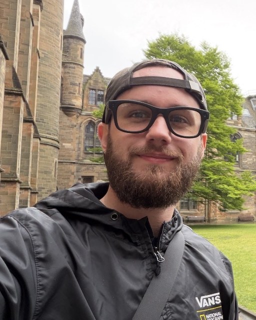 Dylan Hughes, wearing glasses and a backwards cap, outside a stone building.